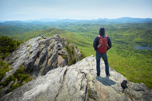 A hiker stands on a rocky ledge, overlooking a lush green valley and distant mountains under a clear sky.