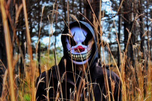 A figure in a dark hoodie stands among tall grass, wearing a creepy clown mask in a forested setting.
