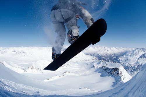A snowboarder jumps off a snowy peak, with a clear blue sky and mountain range in the background.