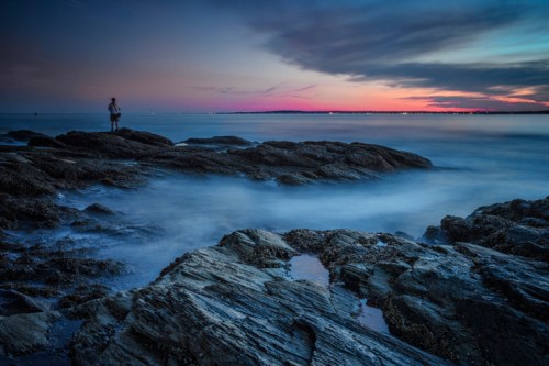 A serene coastal scene at dusk, featuring smooth rocks and a person standing by the water, with a colorful sky.
