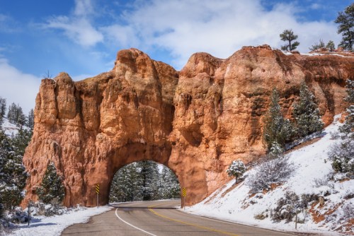 A scenic road curves through a large, reddish rock arch, surrounded by snow-covered trees and a blue sky.