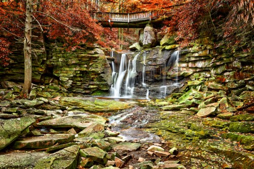 A serene waterfall cascades over mossy rocks, surrounded by vibrant autumn foliage and a wooden bridge above.