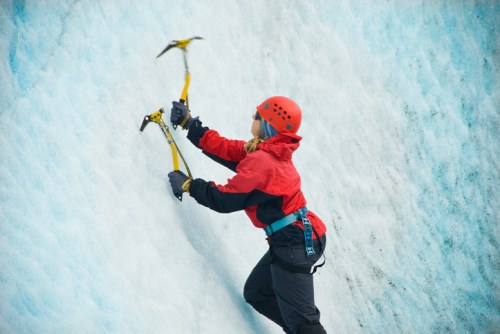A climber in a red jacket and helmet ascends a blue glacier using ice axes.