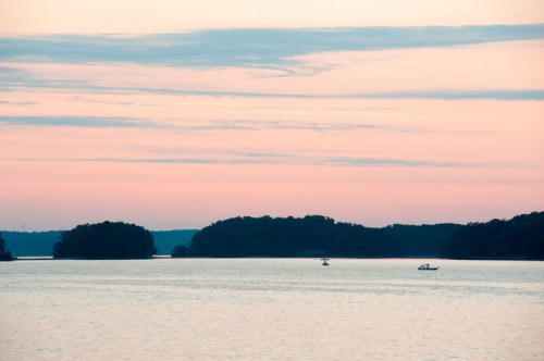 A serene lake at sunset, with soft pink skies and small boats on the water, surrounded by distant islands.
