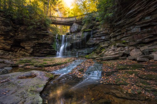 A serene waterfall cascades over rocks, surrounded by lush greenery and autumn leaves, with a wooden bridge above.