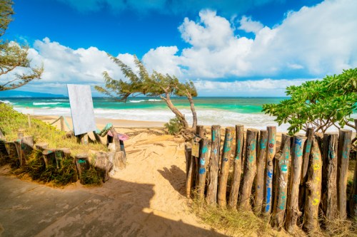 A scenic beach view with turquoise water, sandy shore, and a wooden fence surrounded by trees under a blue sky.
