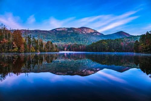 A serene lake reflects colorful autumn trees and mountains under a blue sky with wispy clouds.