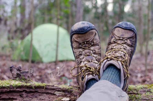 A pair of worn hiking boots resting on a log, with a green tent visible in the blurred background of a forest.