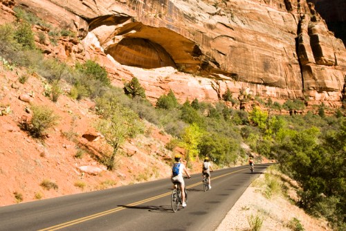 Cyclists ride along a road surrounded by red rock formations and greenery in a scenic canyon landscape.