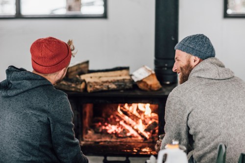 Two men in cozy sweaters and beanies sit by a warm fire, enjoying each other's company in a rustic setting.