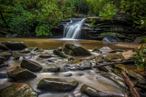 A serene waterfall cascades over rocks into a calm pool, surrounded by lush greenery in a tranquil forest setting.
