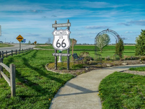 Sign for Historical Route 66 in Illinois, surrounded by green fields and a pathway leading to an archway.