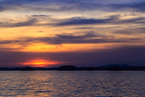 A serene sunset over a calm lake, with colorful clouds reflecting on the water and distant mountains in the background.