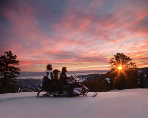 Three people on a snowmobile watch a vibrant sunset over a snowy landscape with trees in the foreground.