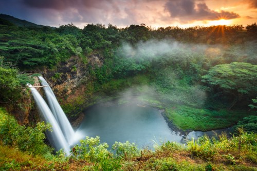A stunning waterfall cascades into a serene pool, surrounded by lush greenery and a colorful sunset sky.