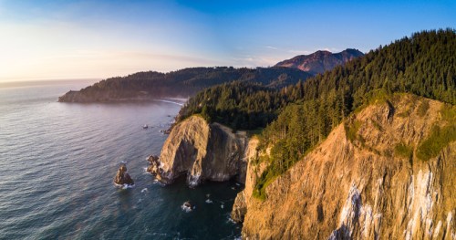 Aerial view of a rugged coastline with cliffs, lush forests, and calm ocean waters under a clear sky.