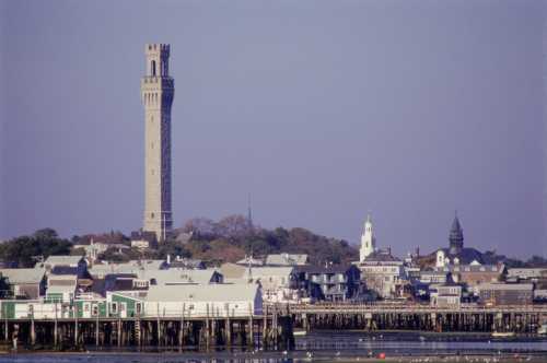 A coastal town with a tall tower, historic buildings, and a pier reflecting in the water under a clear sky.