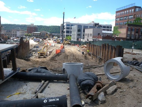 Construction site with heavy machinery, dirt, and materials, surrounded by buildings and a clear blue sky.