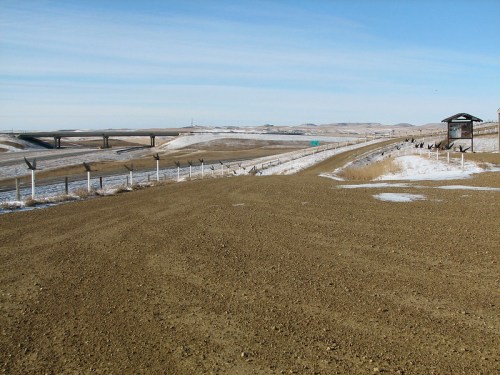 A gravel road leads to a snowy landscape with a highway and overpass in the distance under a clear blue sky.