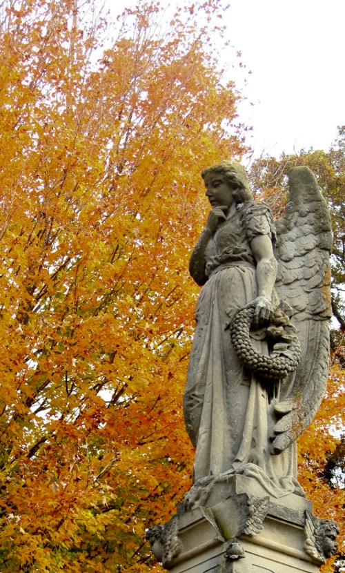 A stone angel statue holding a wreath, set against a backdrop of vibrant autumn leaves.