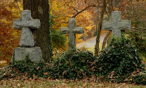 Three weathered stone crosses surrounded by ivy and fallen leaves, set against a backdrop of autumn trees.