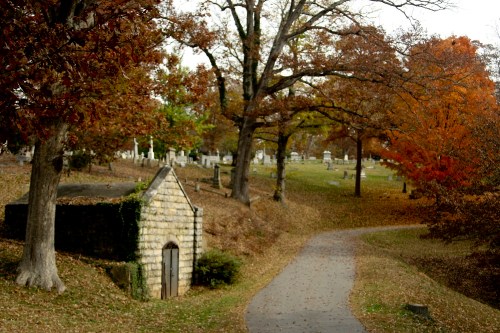 A winding path through a cemetery surrounded by autumn trees and a small stone mausoleum.