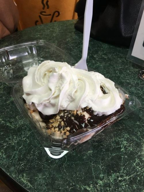 A dessert in a clear container topped with whipped cream, chocolate sauce, and nuts, sitting on a green table.