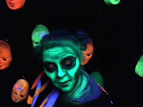 A person with glowing green face paint and dark eyes stands among eerie, illuminated masks in a dark setting.