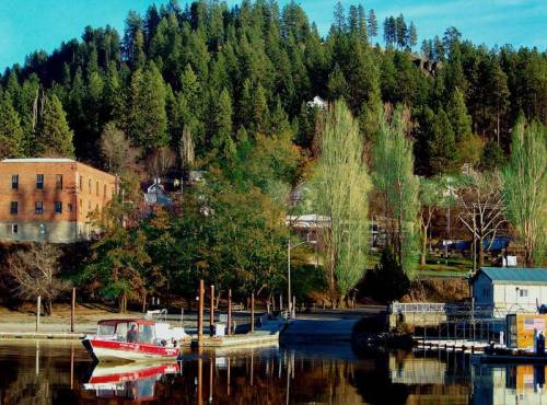 A serene lakeside scene with a red boat, green trees, and a backdrop of hills and buildings reflecting in the water.
