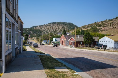 A quiet street in a small town, lined with buildings and hills in the background under a clear blue sky.