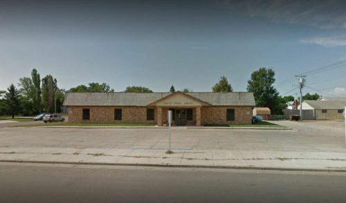 A stone building with a sloped roof, labeled "Public Library," surrounded by trees and a paved area.