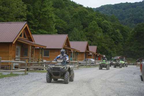 ATVs ride past log cabins in a wooded area, with mountains in the background under a cloudy sky.