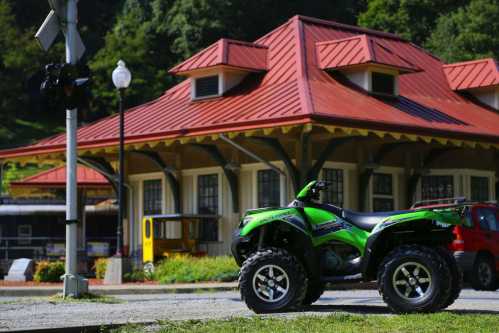 A green ATV parked near a building with a red roof and train tracks in a scenic outdoor setting.
