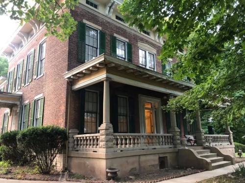 A historic brick house with green shutters, a porch, and surrounded by trees and shrubs.