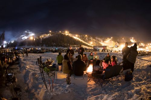 A group of people gathered around a campfire in the snow, with a ski resort illuminated in the background at night.