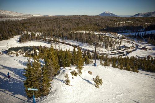 A snowy landscape with a ski resort, pine trees, and mountains in the background under a clear blue sky.