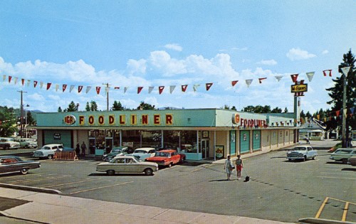 A vintage grocery store with colorful banners, surrounded by classic cars and two people walking in the parking lot.