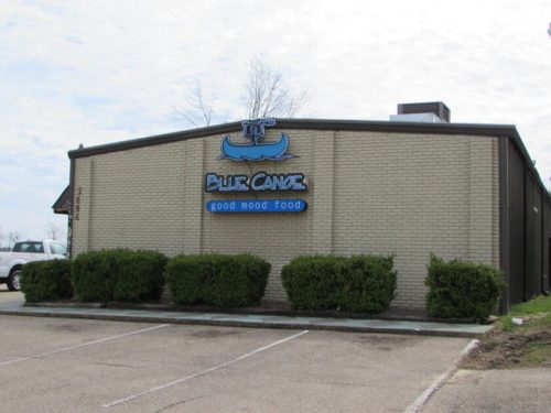 Exterior of a restaurant named "Blue Canoe," featuring a sign that reads "good mood food" and neatly trimmed bushes.