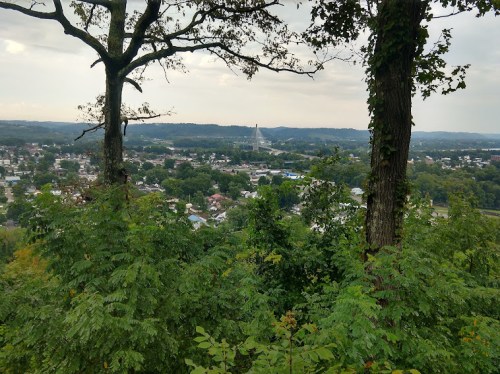 A scenic view of a town nestled in a valley, framed by trees and hills under a cloudy sky.