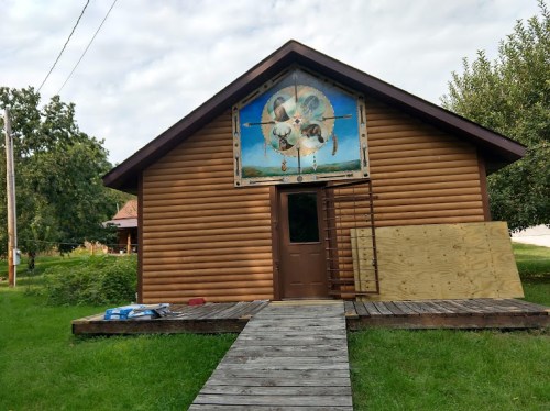 A wooden cabin with a mural on the front, surrounded by grass and trees, featuring a wooden deck leading to the door.
