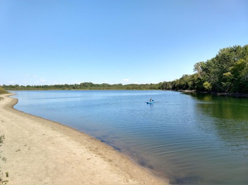 A calm river scene with a sandy shore and a person kayaking in the distance under a clear blue sky.