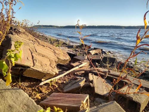 A shoreline with broken bricks and plants, overlooking a calm river under a clear blue sky.