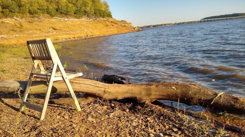 A wooden chair sits on a rocky shore by a calm lake, with trees and a distant pier in the background.