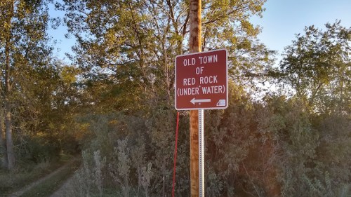 Signpost indicating the direction to the submerged Old Town of Red Rock, surrounded by trees and a dirt path.