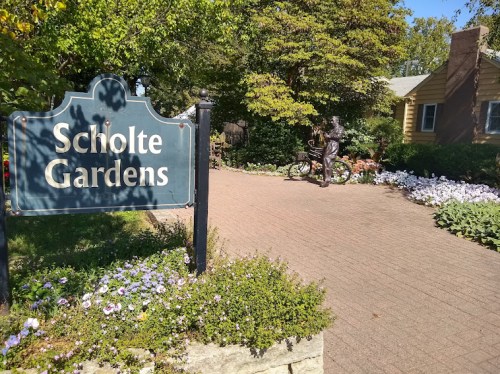 Sign for Scholte Gardens with a pathway, flowers, and a statue of a person near a bicycle.