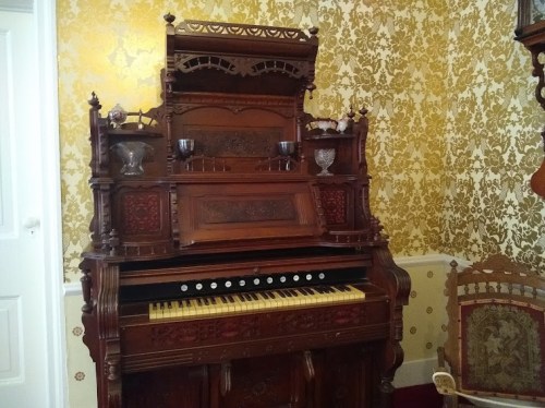 A vintage wooden organ with intricate carvings, set against a patterned yellow wallpaper in a historic room.