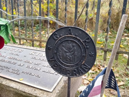 A close-up of a GAR veteran medal hanging near a gravestone, with an American flag and autumn foliage in the background.