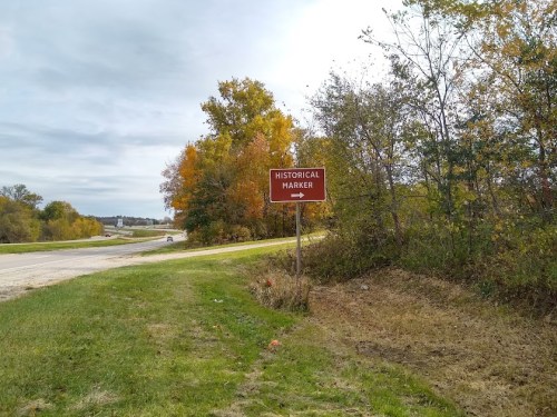 A brown historical marker sign stands beside a road, surrounded by trees with autumn foliage.