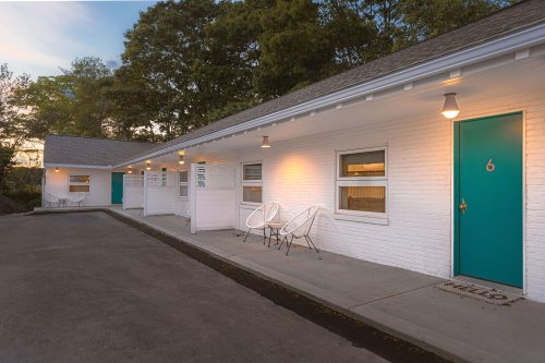 A white motel with teal doors and outdoor seating, surrounded by trees at sunset.