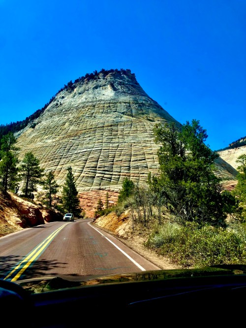 A scenic view of a towering rock formation beside a winding road, surrounded by trees under a clear blue sky.